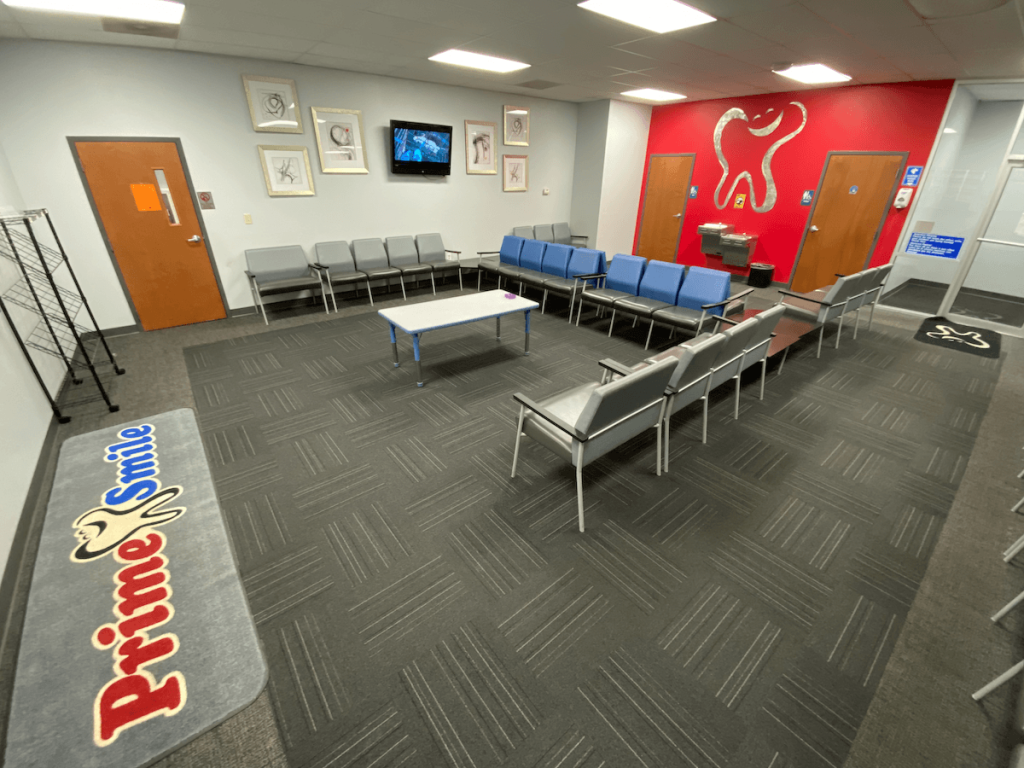 Large patient waiting area with grey and blue chairs surrounding a low center table, featuring carpet flooring and a red Prime Smile wall art logo