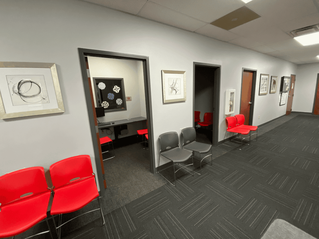 Interior hallway in a medical office lined with grey walls, red waiting room chairs, framed artwork, and private consultation rooms.