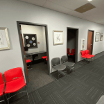 Interior hallway in a medical office lined with grey walls, red waiting room chairs, framed artwork, and private consultation rooms.