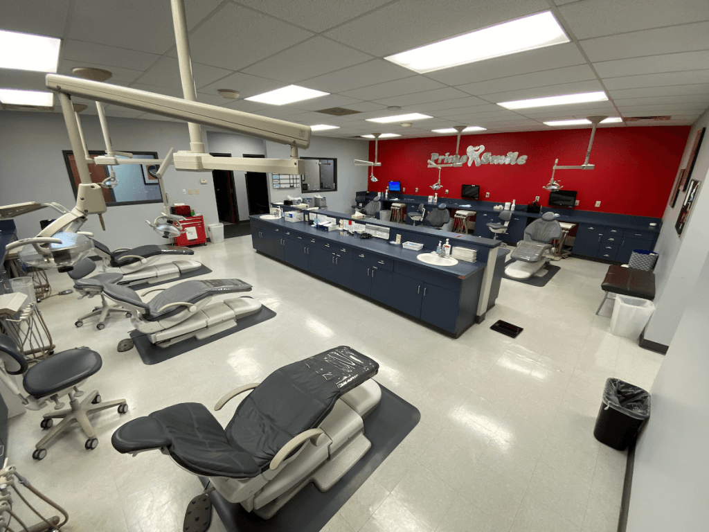 Spacious open-plan dental bay with rows of patient treatment chairs, clinical cabinets, and a red feature wall with the Prime Smile logo