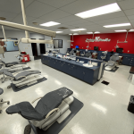Spacious open-plan dental bay with rows of patient treatment chairs, clinical cabinets, and a red feature wall with the Prime Smile logo