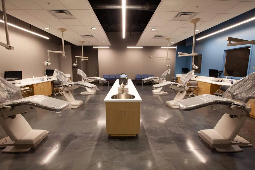 Wide shot of an open bay dental clinic room with multiple patient treatment chairs, overhead operating lights, and central sink stations