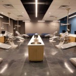 Wide shot of an open bay dental clinic room with multiple patient treatment chairs, overhead operating lights, and central sink stations