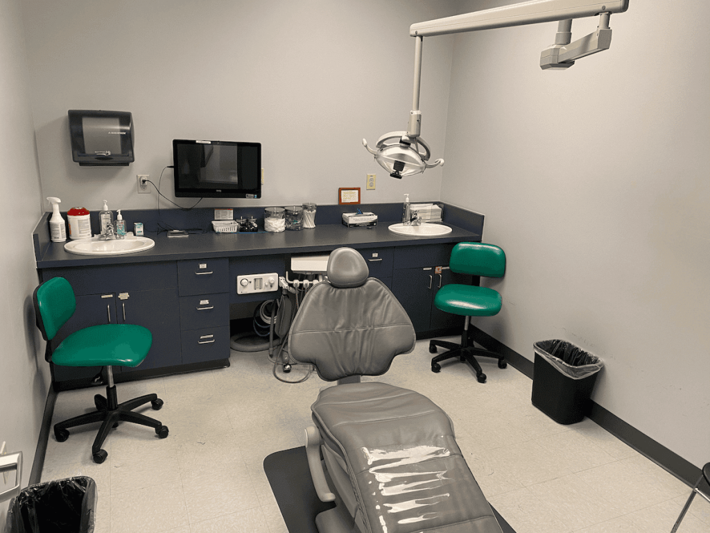 Private dental examination room with a grey patient chair, green operator stools, sink, and overhead dental lighting
