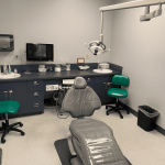 Private dental examination room with a grey patient chair, green operator stools, sink, and overhead dental lighting